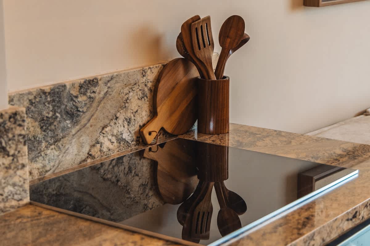 A close-up of a modern kitchen countertop made of light brown granite with dark veining, featuring a sleek black induction cooktop. A wooden utensil holder with various wooden cooking tools and a small wooden cutting board are visible on the counter.