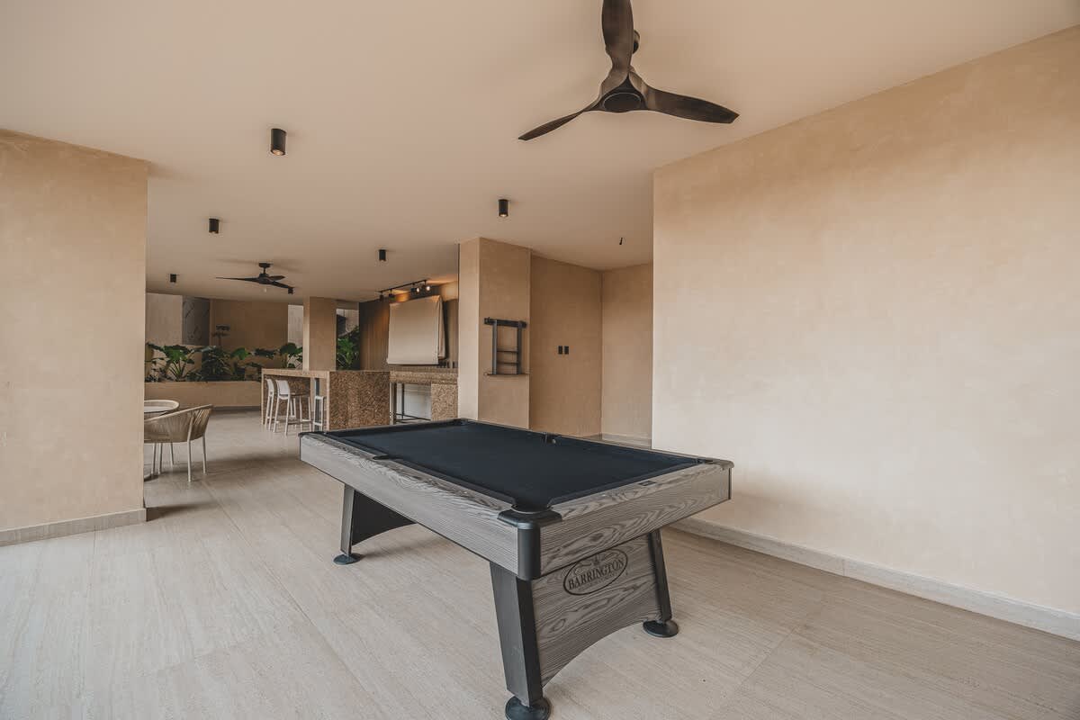 A spacious indoor game area with light-colored tiled flooring, featuring a dark wood pool table with a black felt surface, under a high ceiling with a dark ceiling fan and recessed lighting.