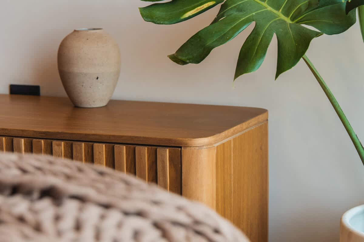 A detailed shot of a modern wooden nightstand with a rounded edge and slatted front, topped with a textured beige vase, next to a cozy knitted blanket.