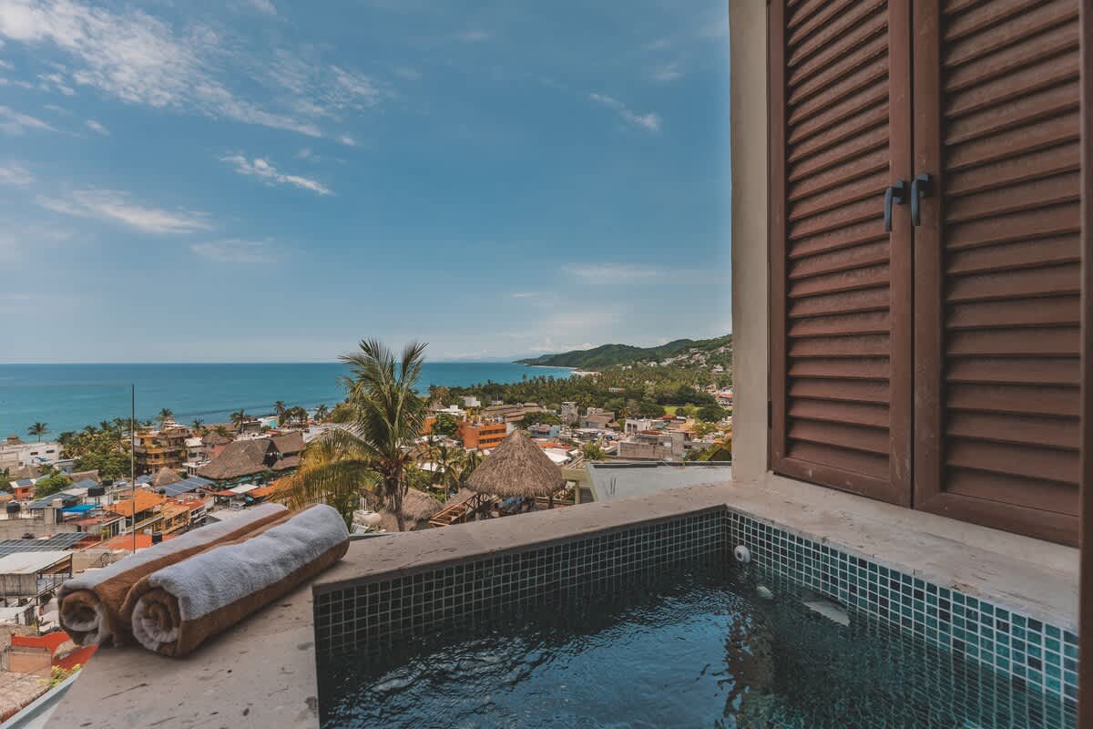 An outdoor hot tub with blue mosaic tiles sits on a balcony, offering panoramic views of the ocean and a coastal town. Rolled white towels are placed on the ledge next to the tub.