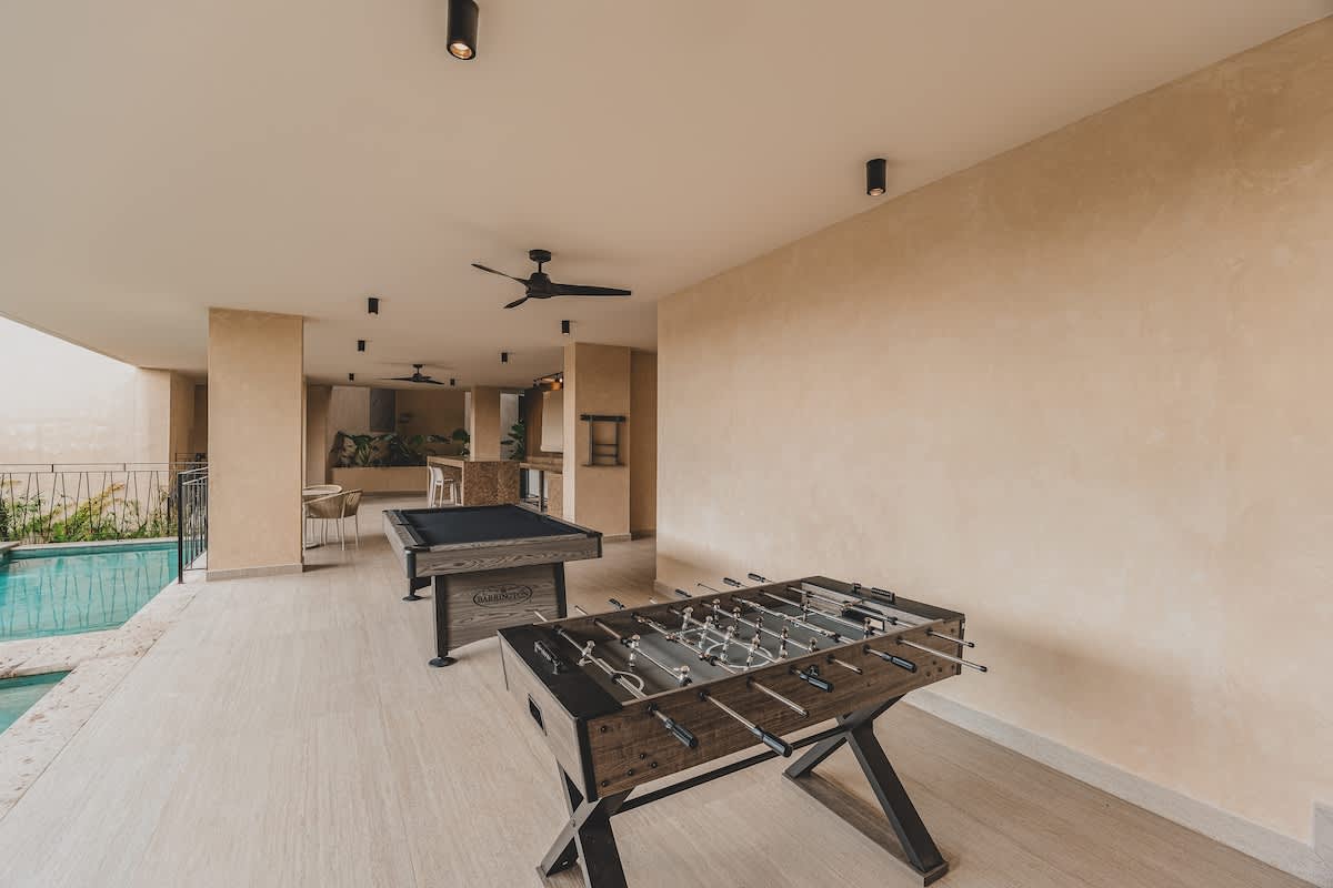 Covered outdoor game area featuring a modern pool table with a black felt surface and a rustic foosball table, set on light-colored tiled flooring with recessed lighting and ceiling fans.