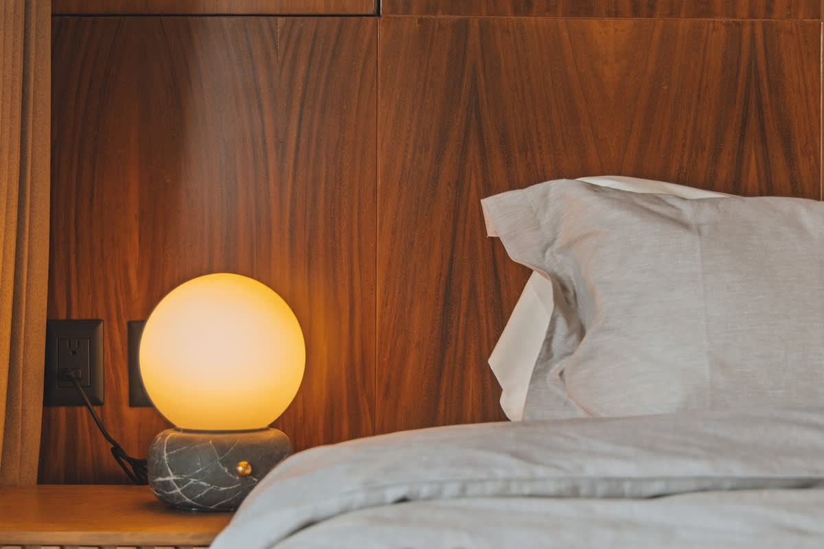 A cozy bedroom features a light gray duvet and pillows, a warm glowing spherical bedside lamp on a marble base, and a rich wooden headboard and wall paneling.