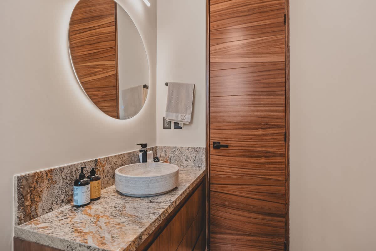 A contemporary bathroom vanity with a light-colored stone vessel sink set on a granite countertop, illuminated by a backlit round mirror, next to a dark wooden door.