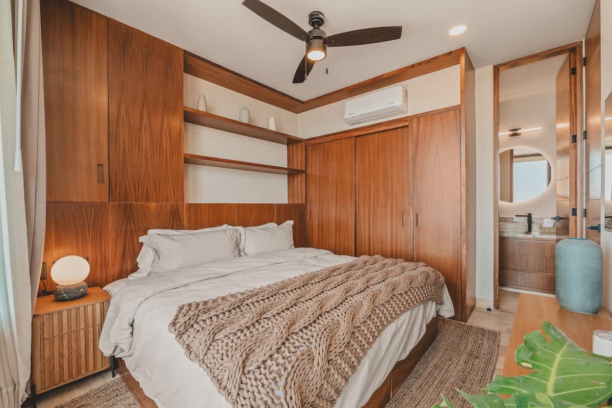 A modern bedroom features a large bed with a beige throw, flanked by a wooden nightstand and lamp. Built-in dark wood cabinetry and open shelving line the walls, with a ceiling fan and AC unit overhead.