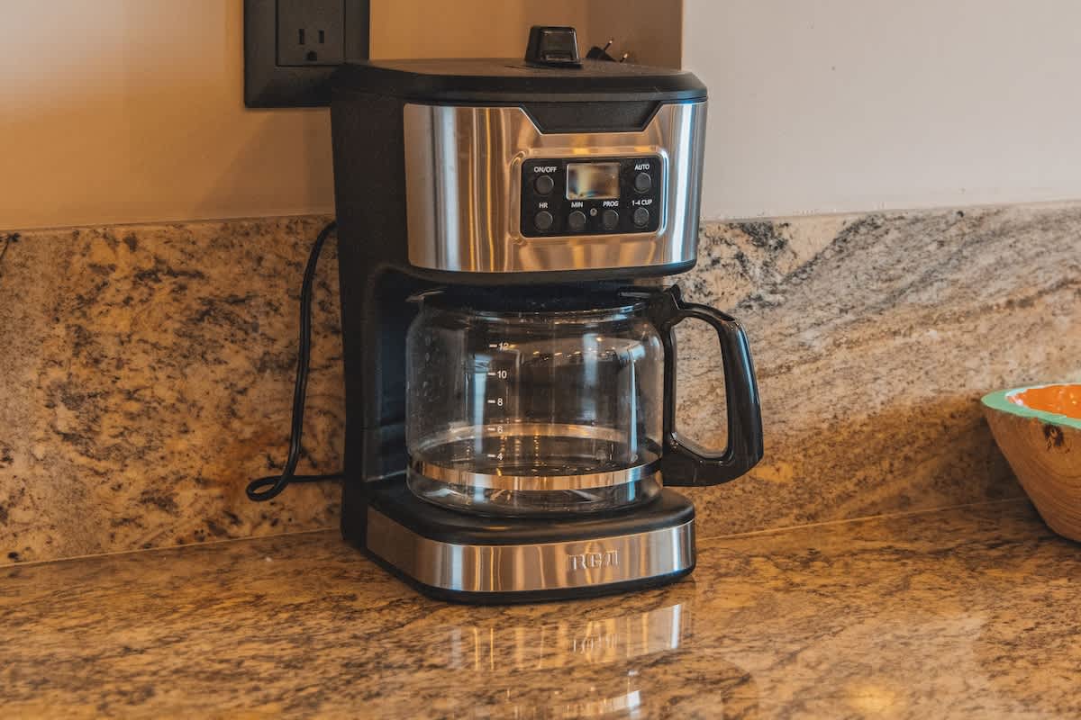 A stainless steel and black coffee maker with a glass carafe sits on a speckled granite countertop, ready for use, against a light-colored wall with an electrical outlet.