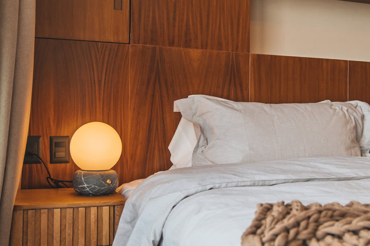 A comfortable bedroom showcases a bed with light gray linens, a spherical bedside lamp emitting a warm glow, and a prominent wooden headboard and wall paneling.