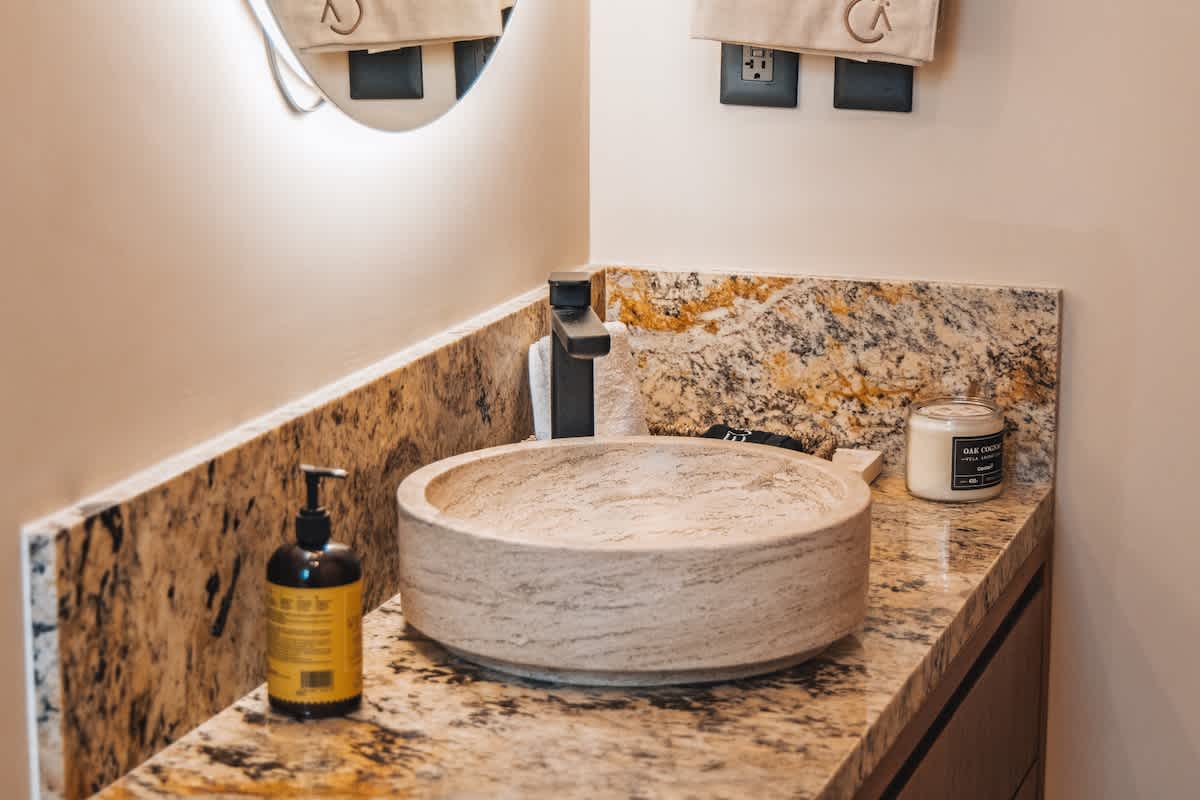 A contemporary bathroom vanity featuring a round stone vessel sink, a matte black faucet, a pump bottle, and a white candle, all resting on a speckled granite countertop with a matching backsplash.