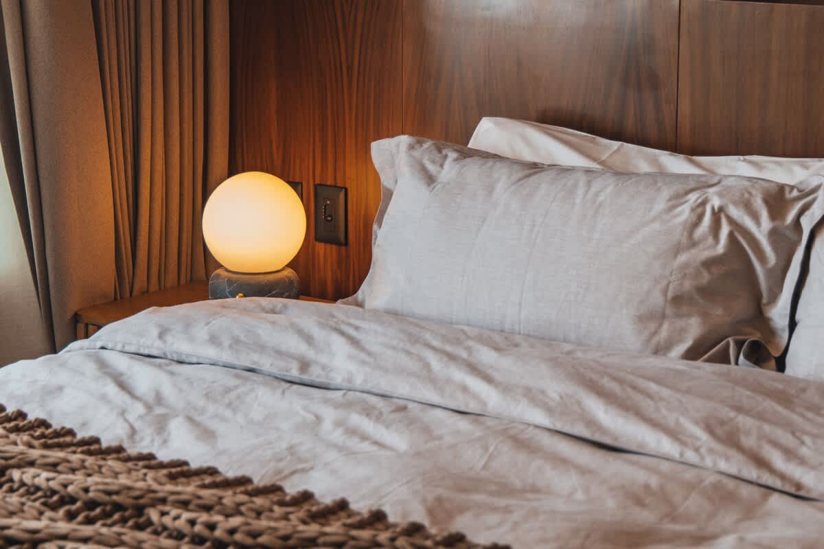 An inviting bedroom features a bed dressed in light gray linens, a warm glowing spherical lamp on a nightstand, and a textured brown throw blanket, all against a wooden paneled wall.