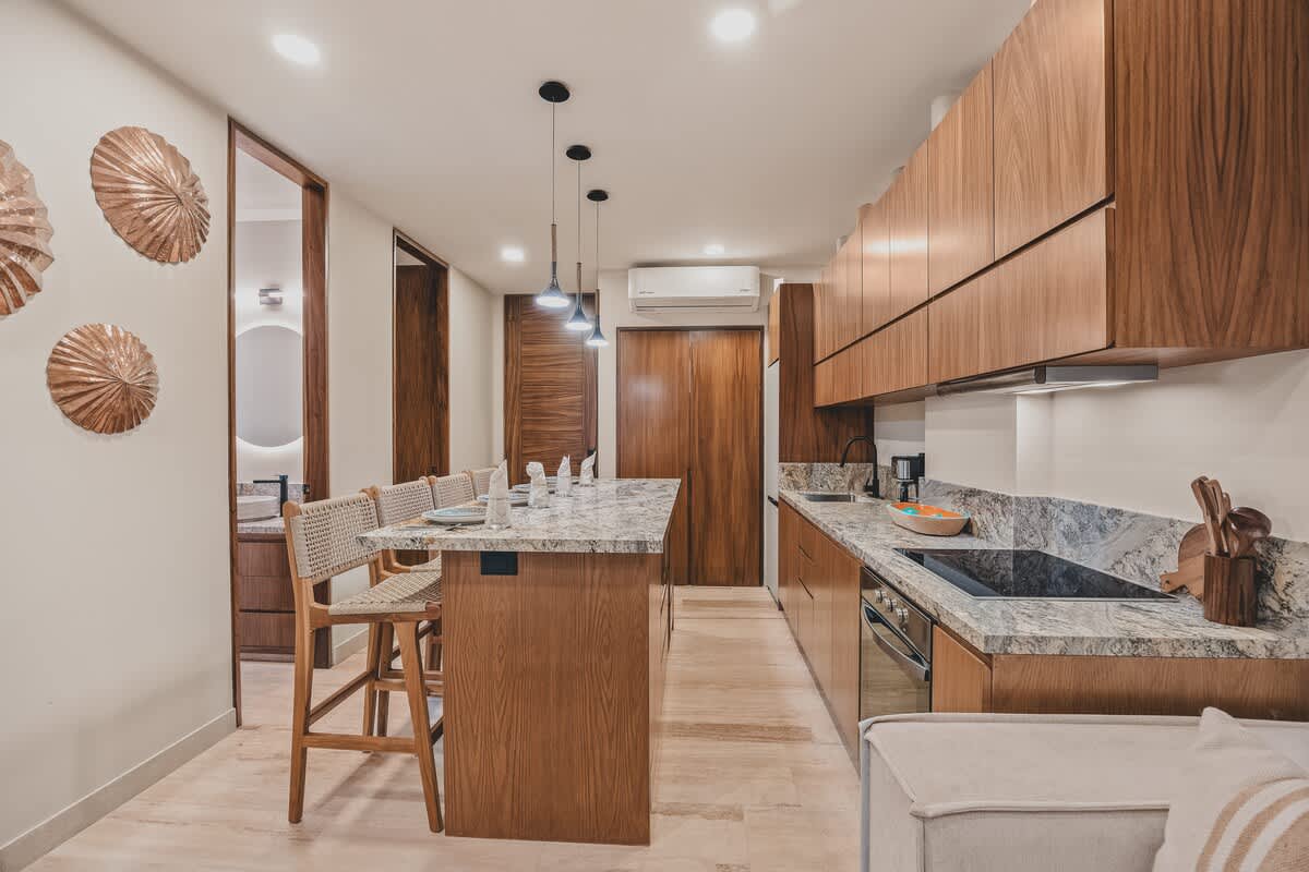 View of the kitchen and dining island from the living space, showcasing the granite countertops, wood cabinetry, and bar seating, with decorative wall art and a modern bathroom entrance.