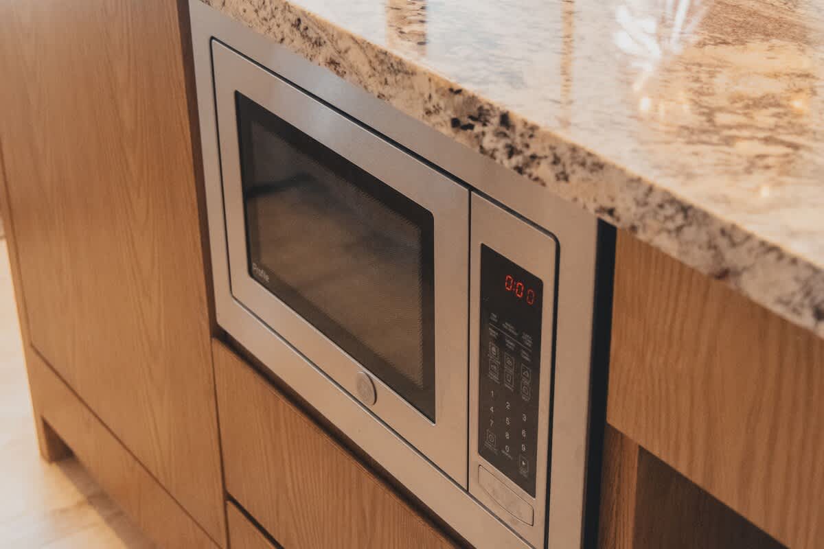 A built-in stainless steel microwave oven with a digital display showing "0:00" and a keypad, integrated seamlessly into light wooden cabinetry beneath a speckled granite countertop.