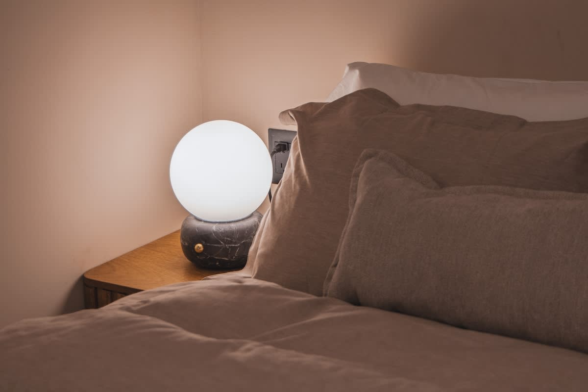 A warm and inviting bedroom corner with a glowing spherical lamp on a wooden nightstand, positioned beside a bed dressed with soft, neutral-toned pillows and bedding.