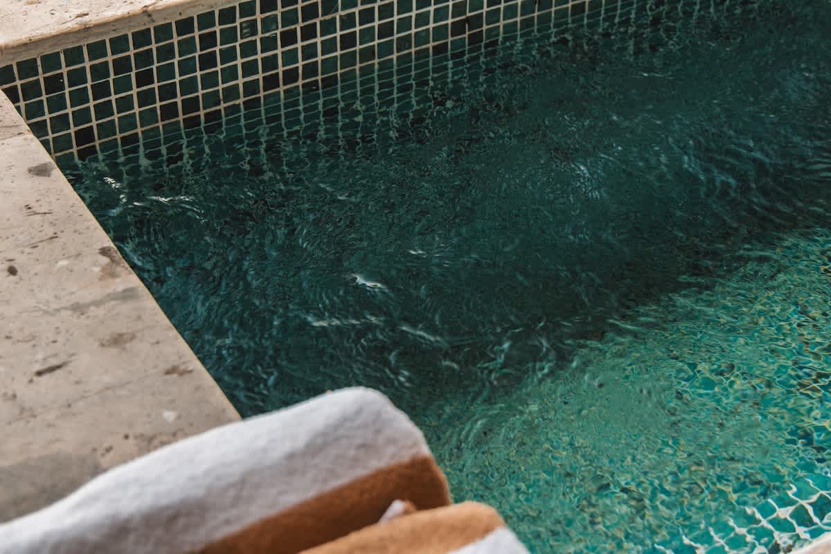 A close-up view of a swimming pool with shimmering turquoise water and dark blue mosaic tiles, bordered by light-colored stone coping and a striped towel.
