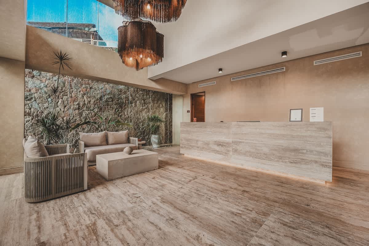 A contemporary lobby with light stone flooring and walls, featuring an illuminated reception desk made of light-colored stone. A seating area with beige sofas and a concrete coffee table is visible, positioned against a textured stone wall with green plants.