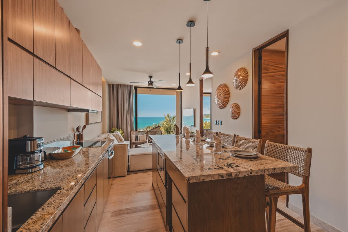 Contemporary kitchen with light wood cabinets, granite countertops, and a dining island with bar stools, seamlessly connecting to a living area with panoramic ocean views.