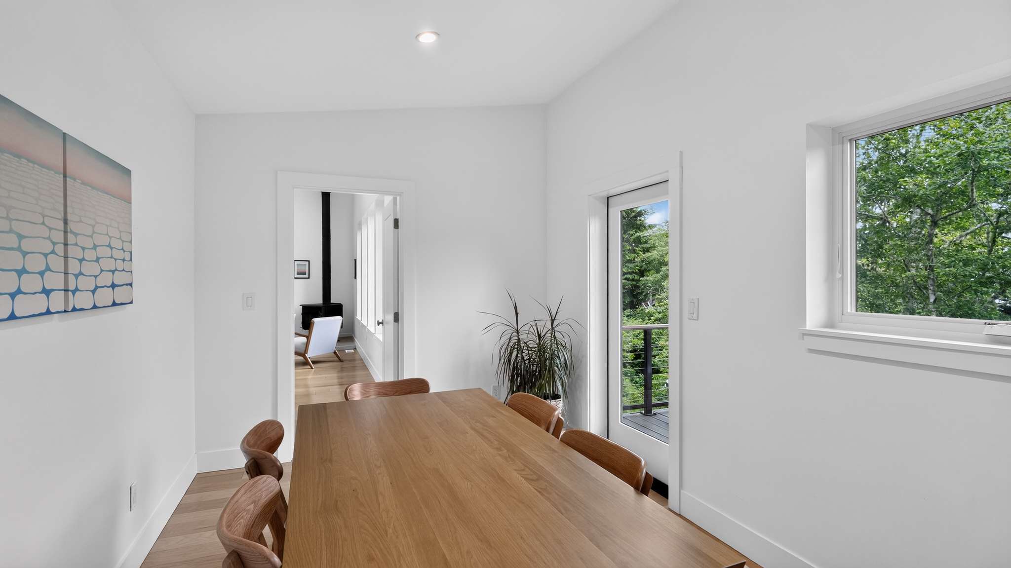 A bright dining area features a long wooden table with six curved wooden chairs, set on light hardwood floors. A glass door opens to a balcony with lush green trees, and a doorway leads to an adjacent room with a modern black fireplace.