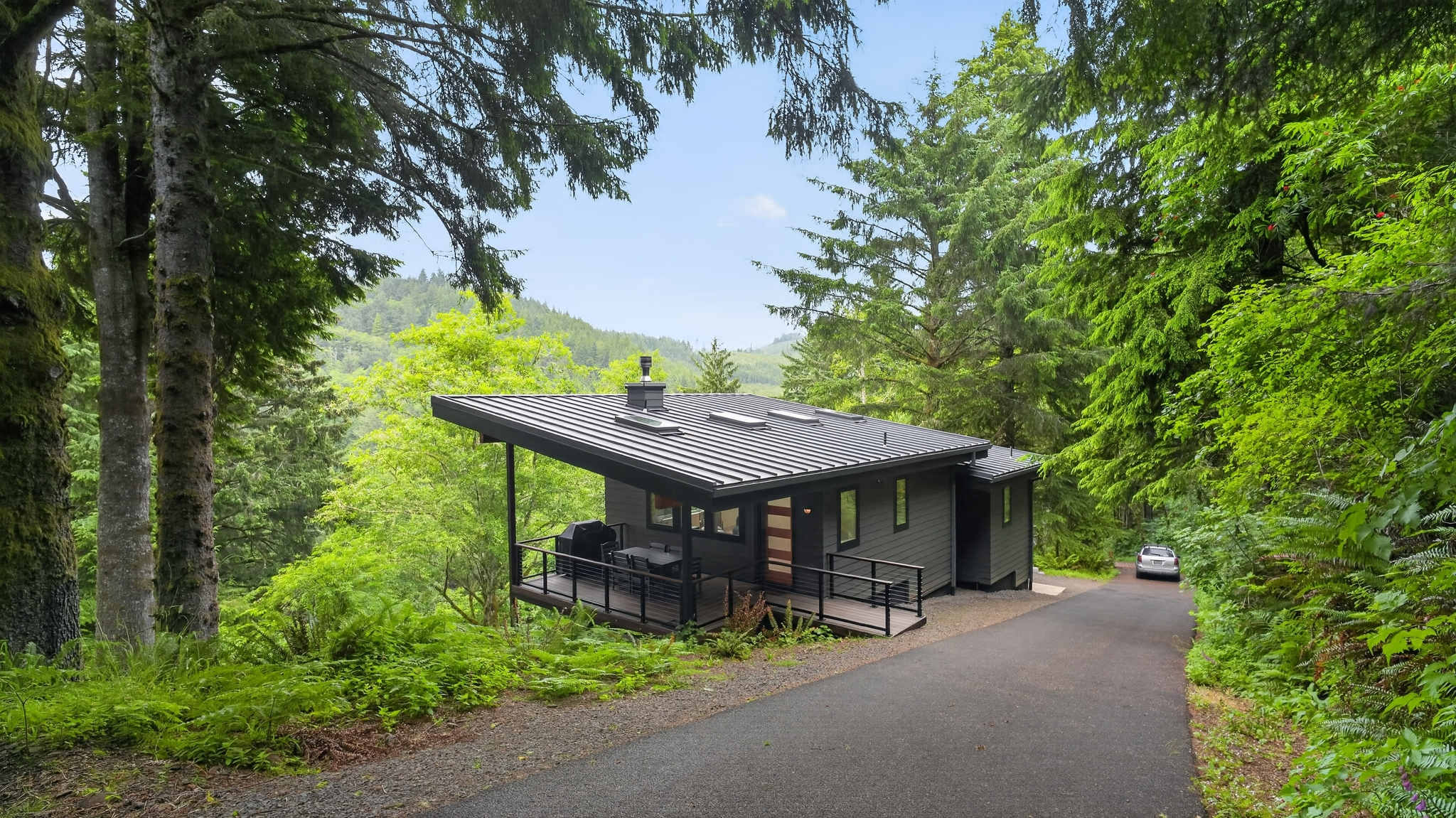 A modern, dark gray house with a sloped metal roof and a covered deck is situated beside a curving asphalt driveway. The property is surrounded by tall, lush evergreen trees under a clear sky.