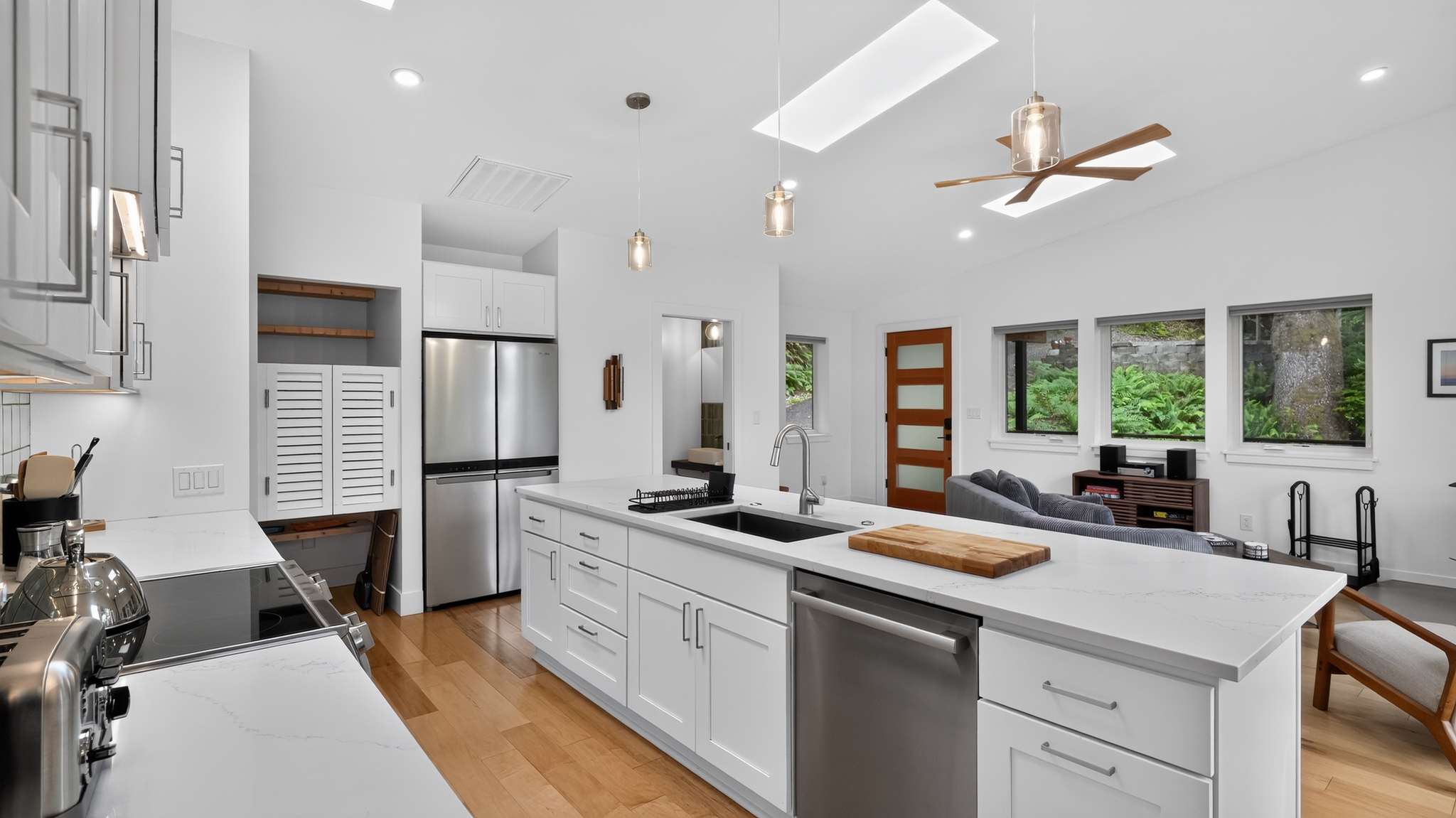 Open-concept kitchen and living area with white walls, light hardwood floors, and a large kitchen island with a black sink. Stainless steel appliances, including a refrigerator and dishwasher, are integrated. A wooden ceiling fan and skylight are visible in the living space.
