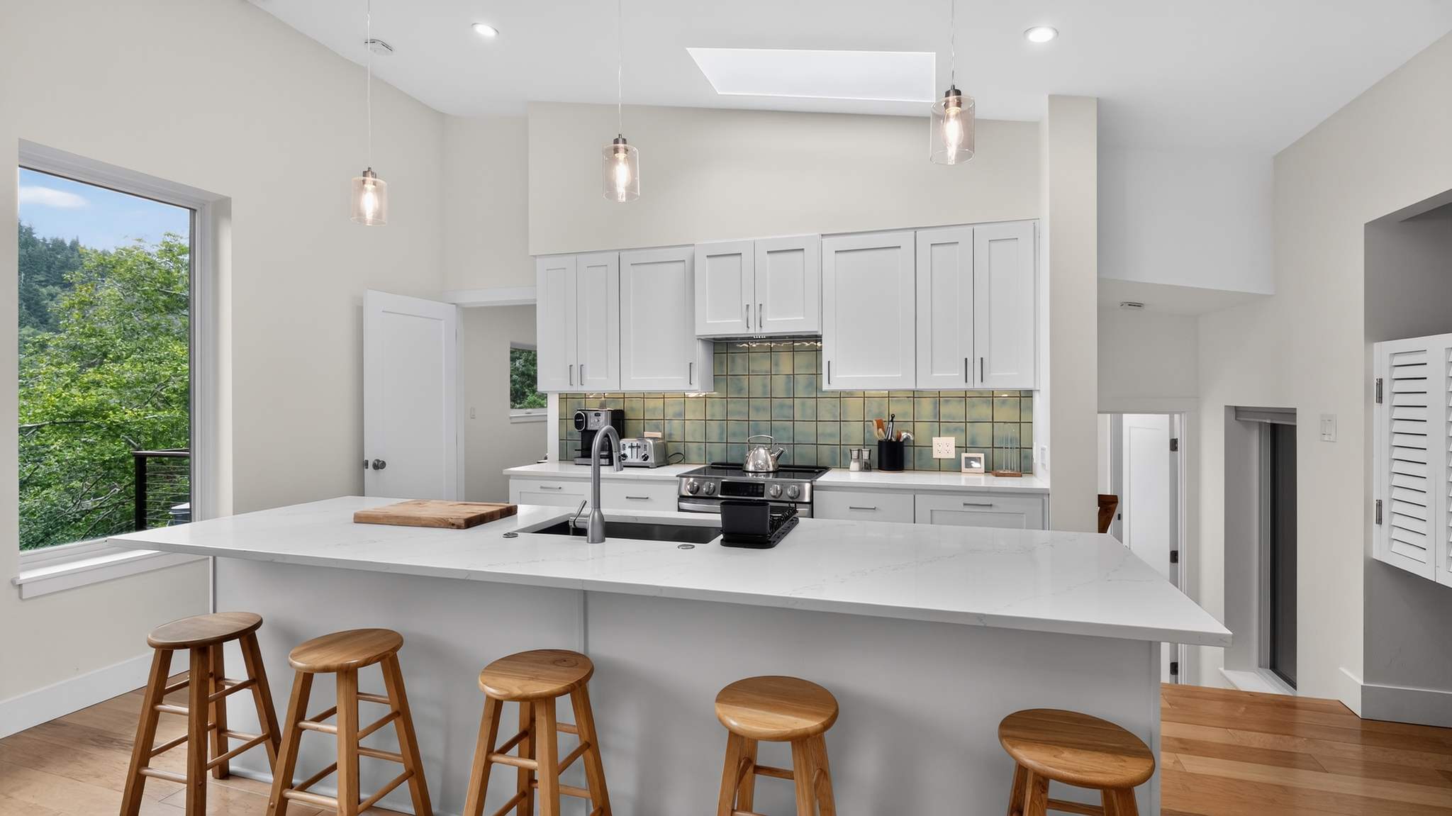 Spacious kitchen island with white quartz countertop and four wooden bar stools, facing a modern kitchen with white shaker cabinets and a green tiled backsplash. Pendant lights hang above the island, and a large window is visible on the left.