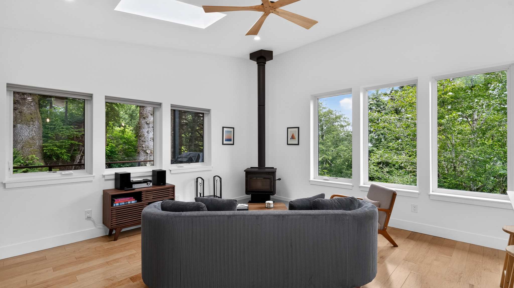 A cozy living room with light hardwood floors and white walls features a dark gray curved sofa facing a modern black wood-burning stove. Multiple windows offer views of the surrounding forest, and a skylight brightens the space.