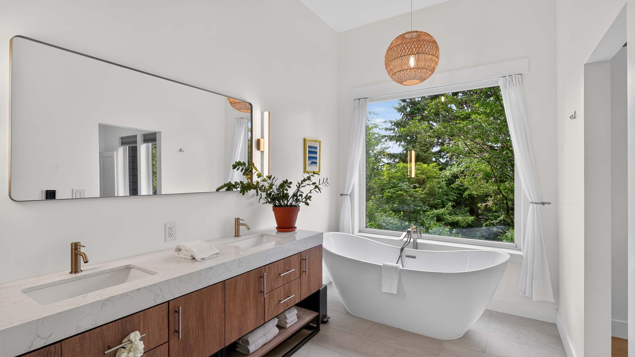Spacious and bright bathroom with a double vanity featuring a wood cabinet and white countertop, a large rectangular mirror, and a freestanding soaking tub by a window with green tree views.