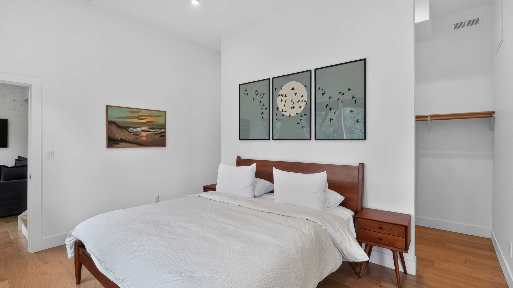 Bedroom with a wooden bed and white bedding, featuring a wooden nightstand and three framed bird artworks above the bed. A painting of a beach scene hangs on an adjacent wall, and a closet entrance is visible on the right.