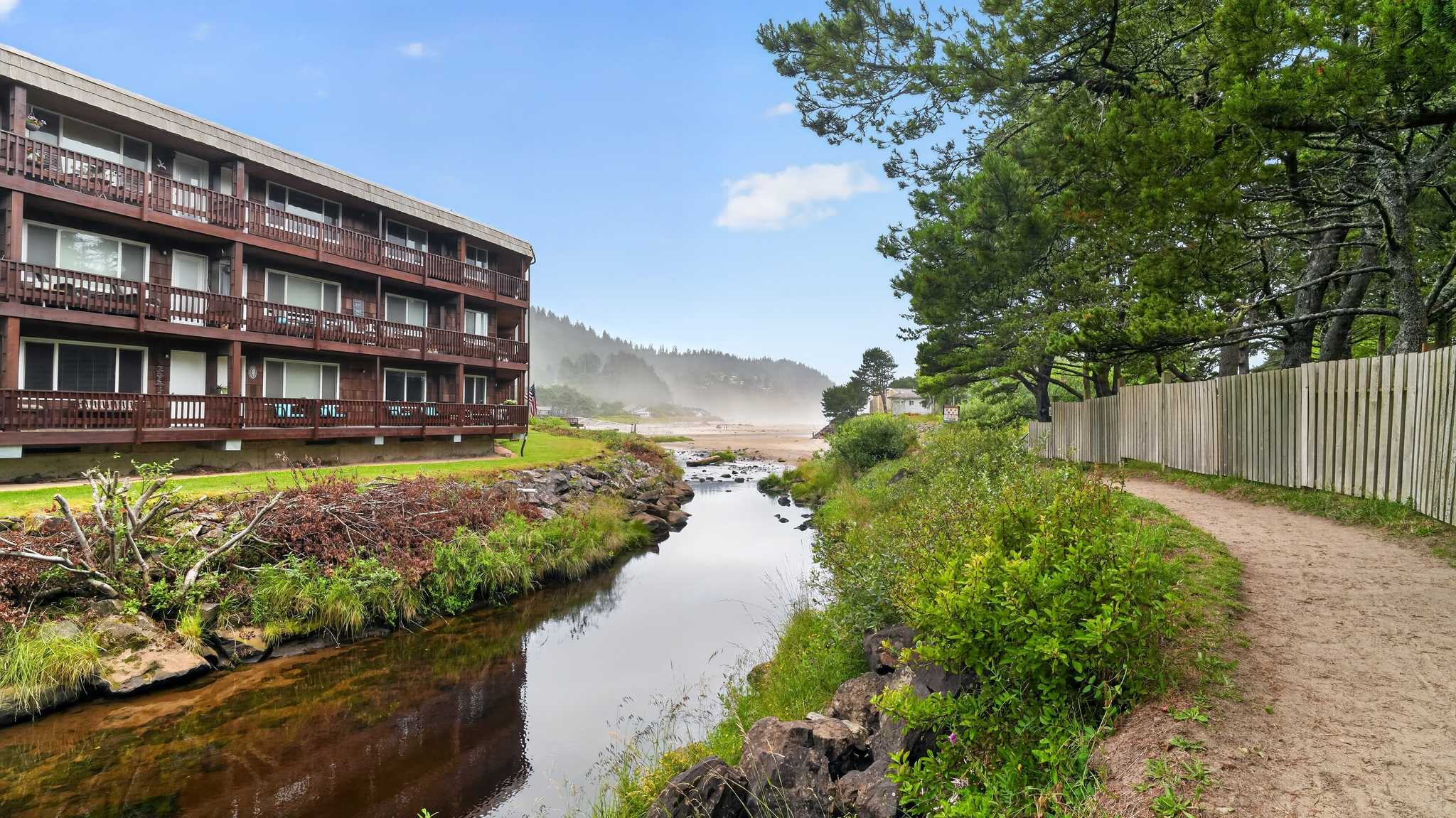 A multi-story wooden building with balconies overlooks a narrow river flowing towards a sandy beach and distant misty hills under a partly cloudy sky. Lush green vegetation lines the riverbanks.
