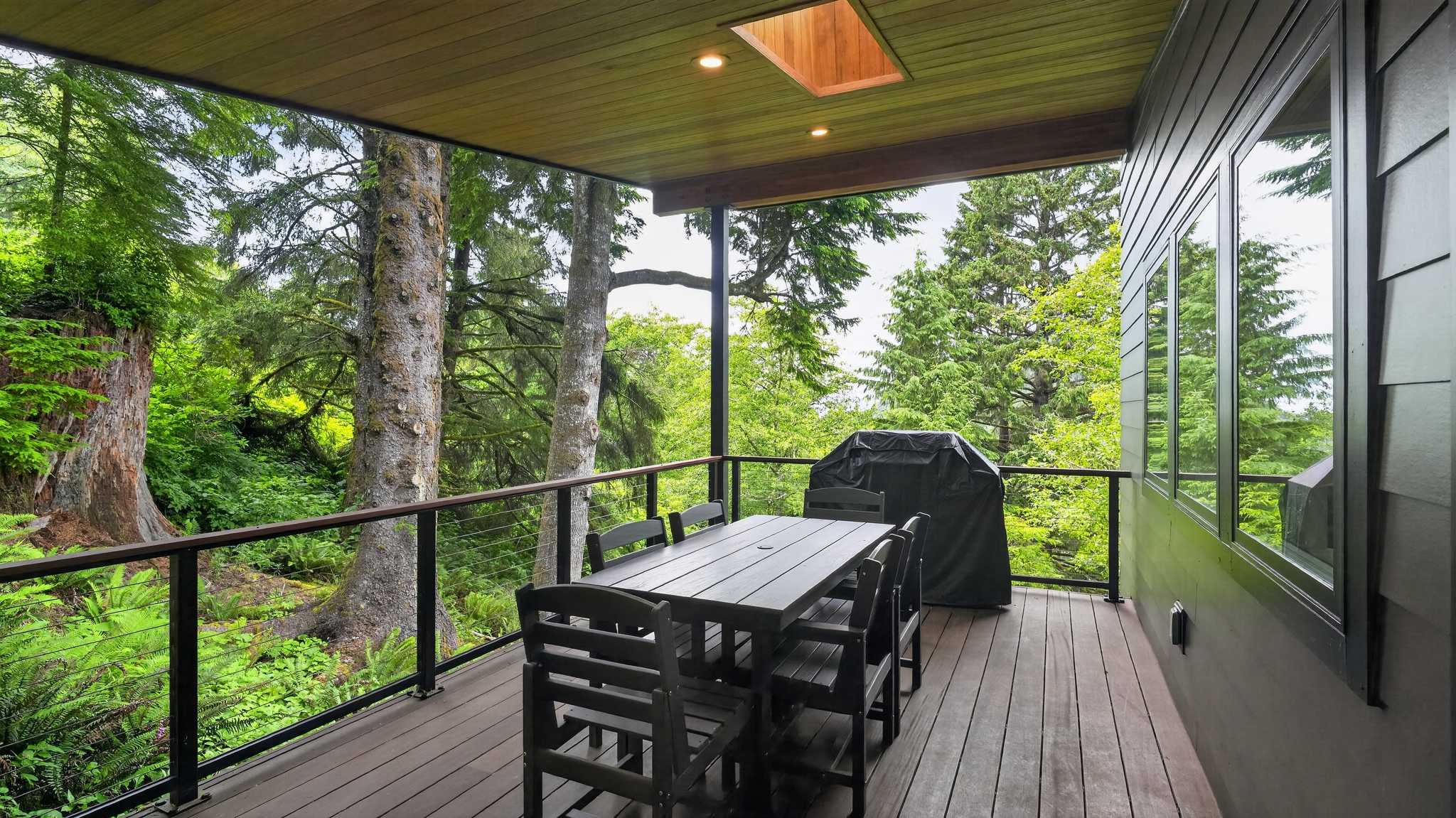 A spacious covered outdoor deck features a dark grey wooden dining table with six matching chairs and a large black grill, all set against a backdrop of dense green forest. The deck has a warm wooden ceiling and black cable railings.