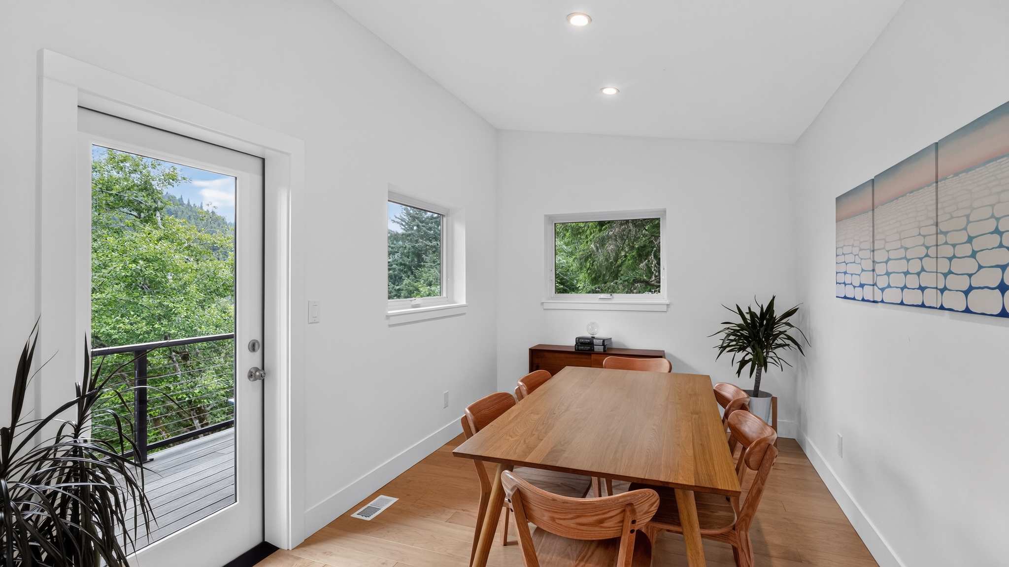 Modern dining room with a long wooden table and matching chairs, white walls, and light wood floors, featuring two windows with tree views and a glass door leading to a balcony.