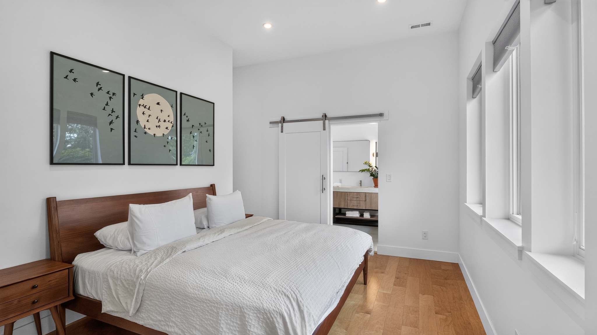 Serene bedroom with a wooden bed frame, white bedding, and two wooden nightstands. Three framed art pieces depicting birds adorn the white wall above the bed, and a barn door leads to an en-suite bathroom.