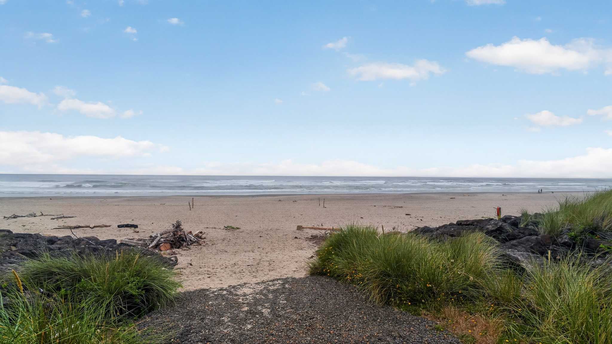 A wide, sandy beach stretches towards the ocean under a partly cloudy blue sky, with gentle waves breaking in the distance. Dark rocks and green dune grasses frame the foreground, leading to a path to the sand.