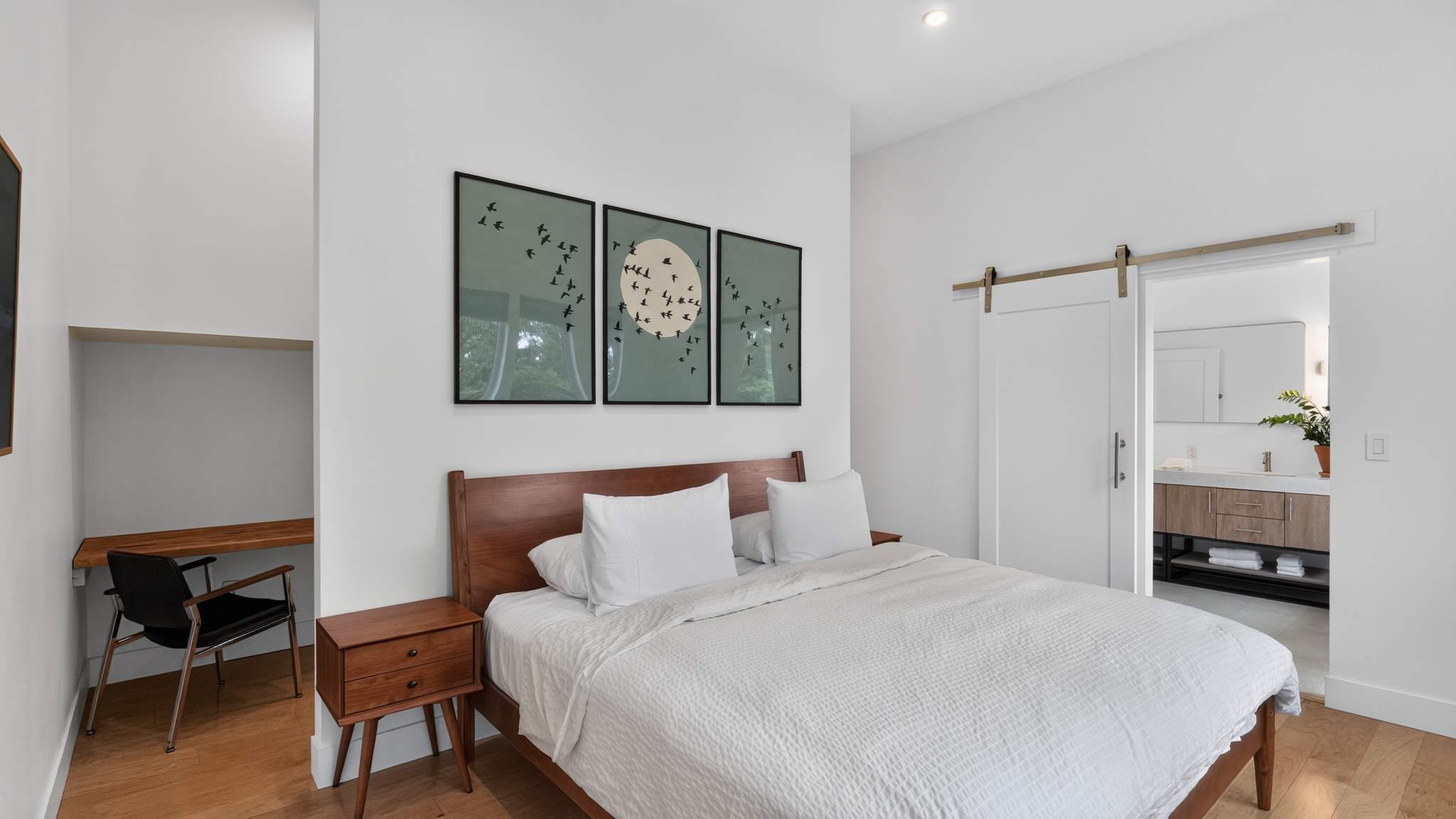 Bedroom featuring a wooden bed with white bedding, flanked by wooden nightstands. A small desk and chair are visible in the corner, and three framed bird artworks hang above the bed. A barn door opens to a bathroom.