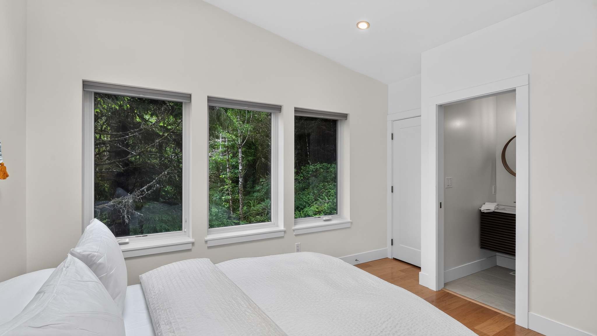 Bright bedroom with white walls and light wood floors, featuring a white bed and three large windows offering immersive views of a lush, dense forest.