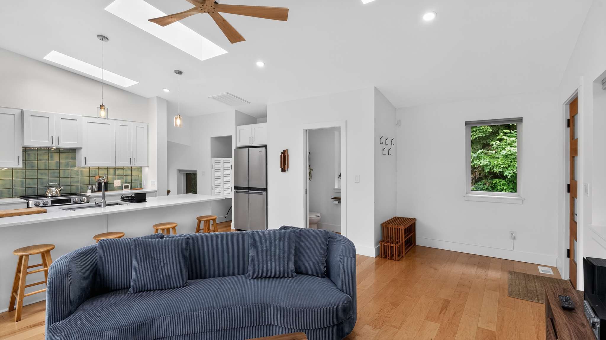 View of the living room from the kitchen, showing a gray curved sofa, hardwood floors, and a glimpse of a separate toilet room, with white walls and natural light.