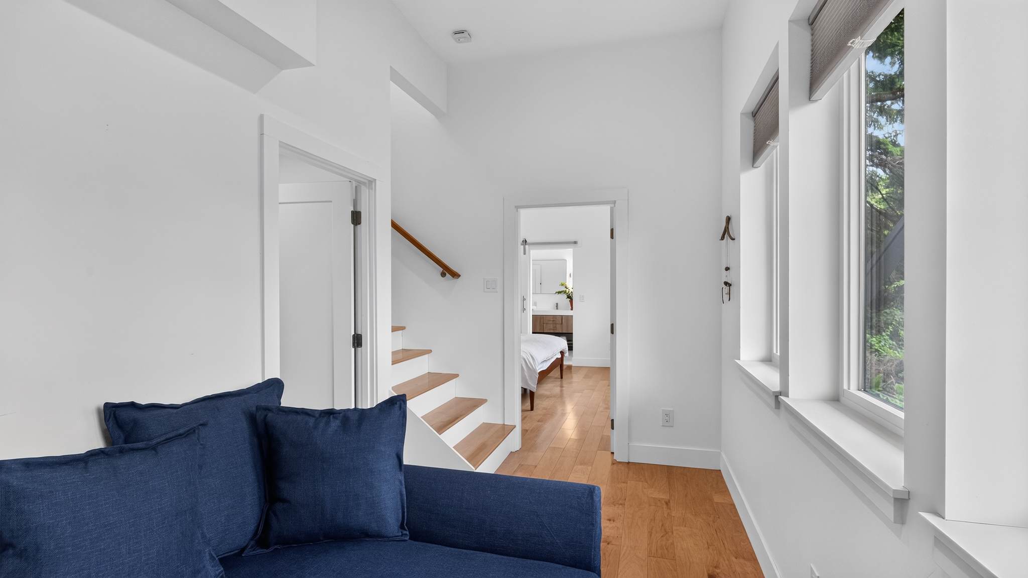 This view of the living space shows the dark blue sofa, light hardwood floors, and a staircase with a wooden handrail. A doorway leads to another room, possibly a bedroom or bathroom, with a glimpse of a bed.