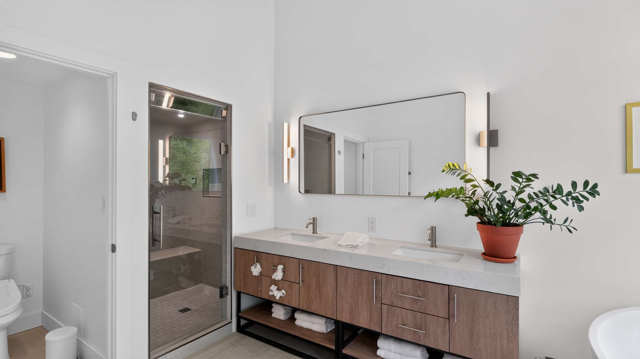 A bright, modern bathroom showcases a double vanity with a light wood cabinet and white countertop, alongside a glass-enclosed shower. A toilet is visible to the left, and a freestanding tub is partially seen on the right.