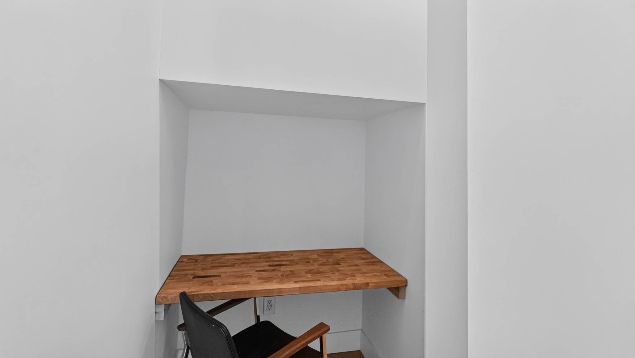 A compact, minimalist office nook features a built-in wooden butcher block desk and a black chair, set within a white alcove. A power outlet is visible on the wall, indicating a functional workspace.