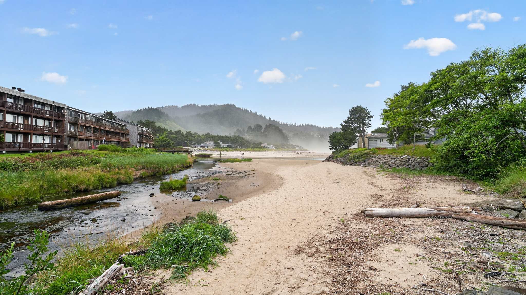 A wide shot of a river flowing into a sandy beach, with a multi-story wooden building on the left and distant misty hills. Logs and rocks are scattered along the riverbanks, and a small white house is visible on the right.