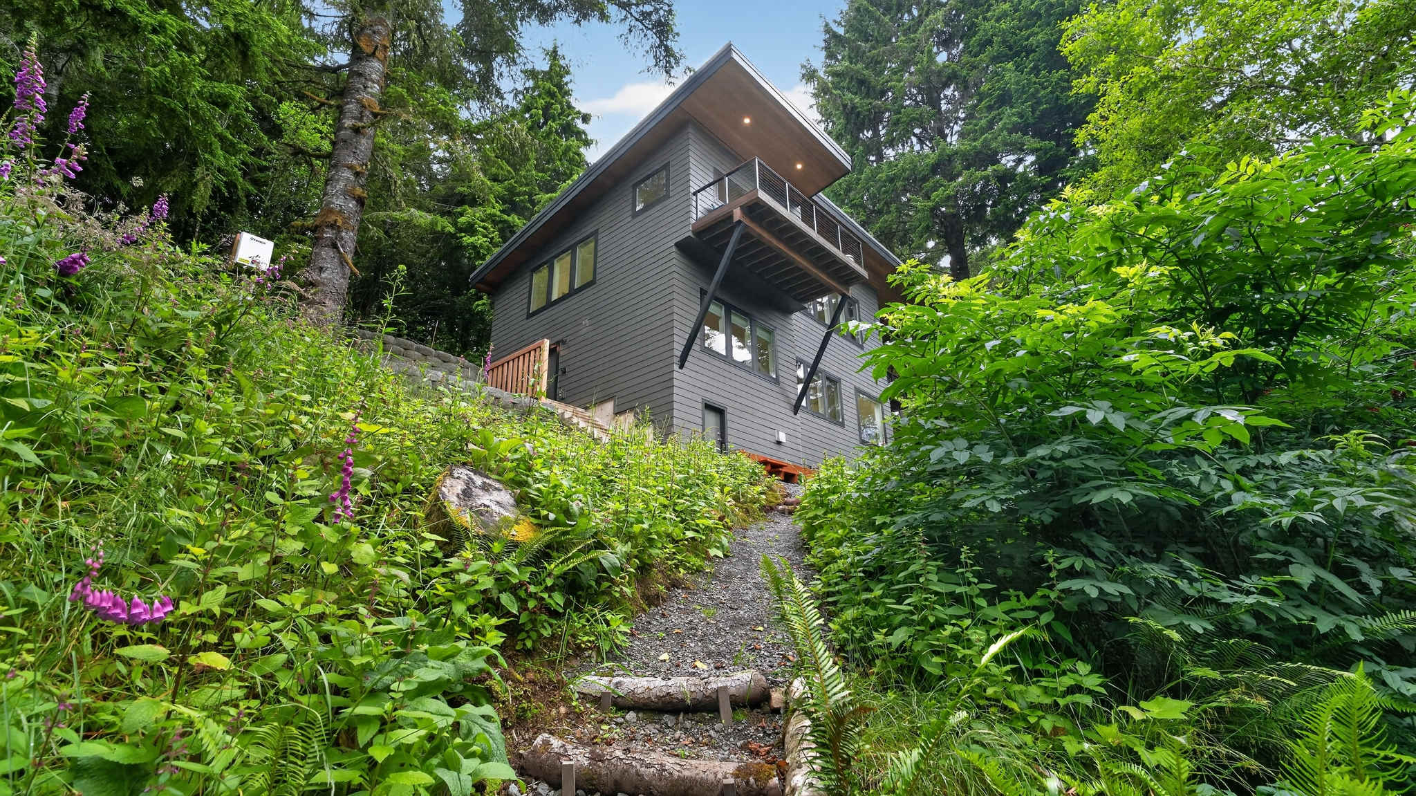 A multi-level modern dark gray house is built into a lush, green hillside, surrounded by dense forest and tall trees. A gravel path with wooden steps leads up to the property, showcasing its elevated position.