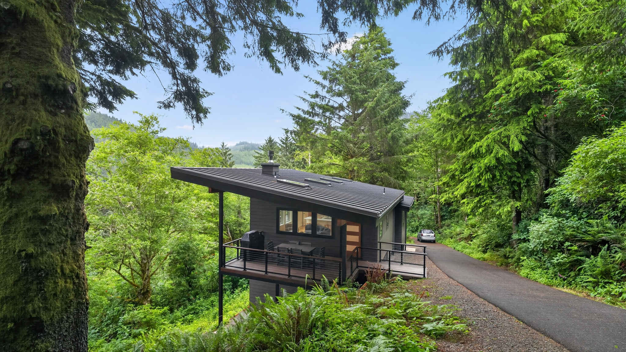 A modern, dark gray house with a prominent sloped metal roof and a spacious deck overlooks a dense forest and distant hills. An asphalt driveway with a parked car curves past the property.