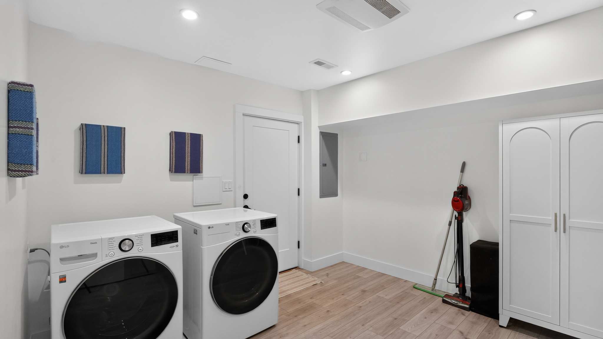 A spacious laundry room features a white front-loading washer and dryer, light wood-look flooring, white walls, and a white storage cabinet. A vacuum cleaner and ironing board are also visible.