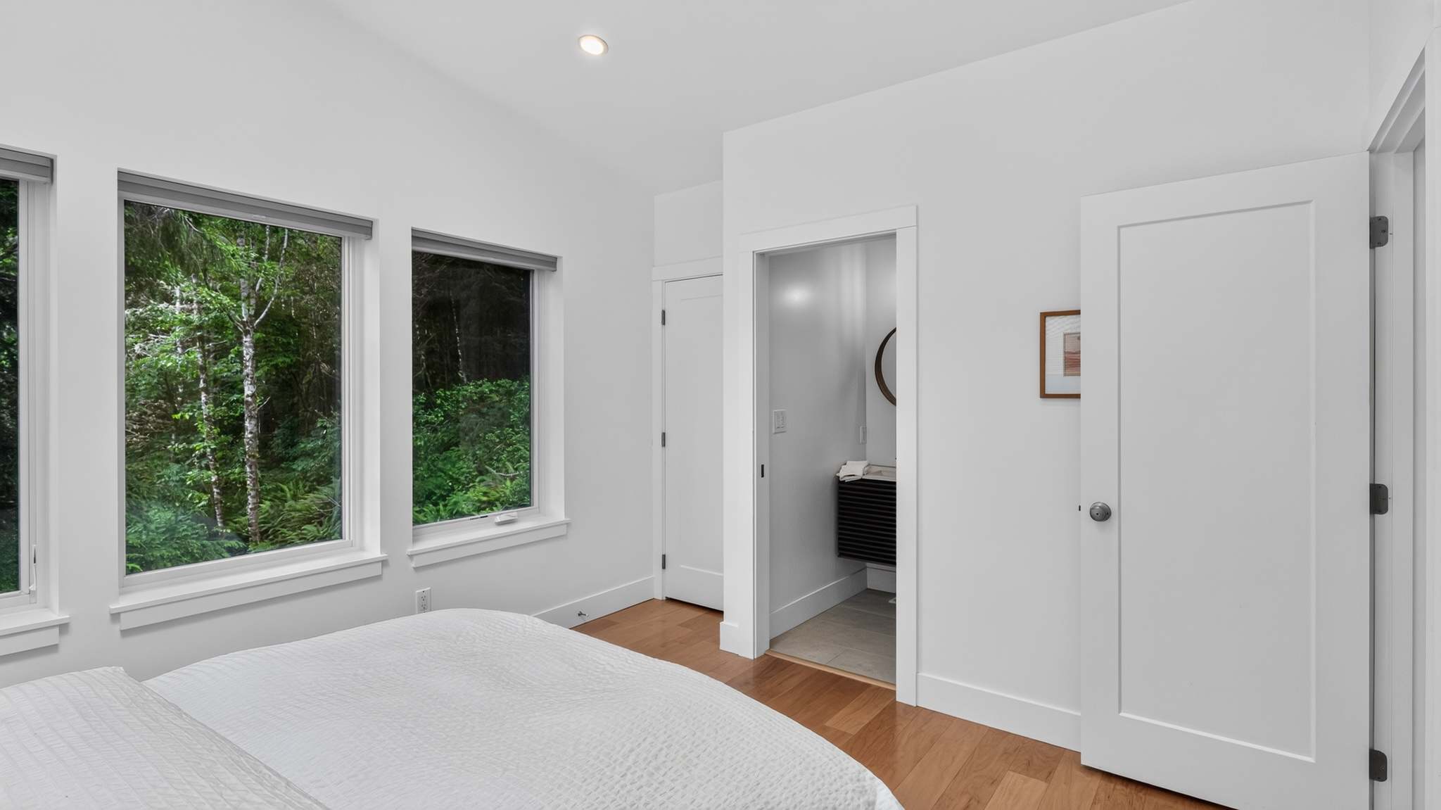 Serene bedroom with white walls, light wood floors, and a white bed, featuring three large windows showcasing dense forest views and a doorway leading to an en-suite bathroom.