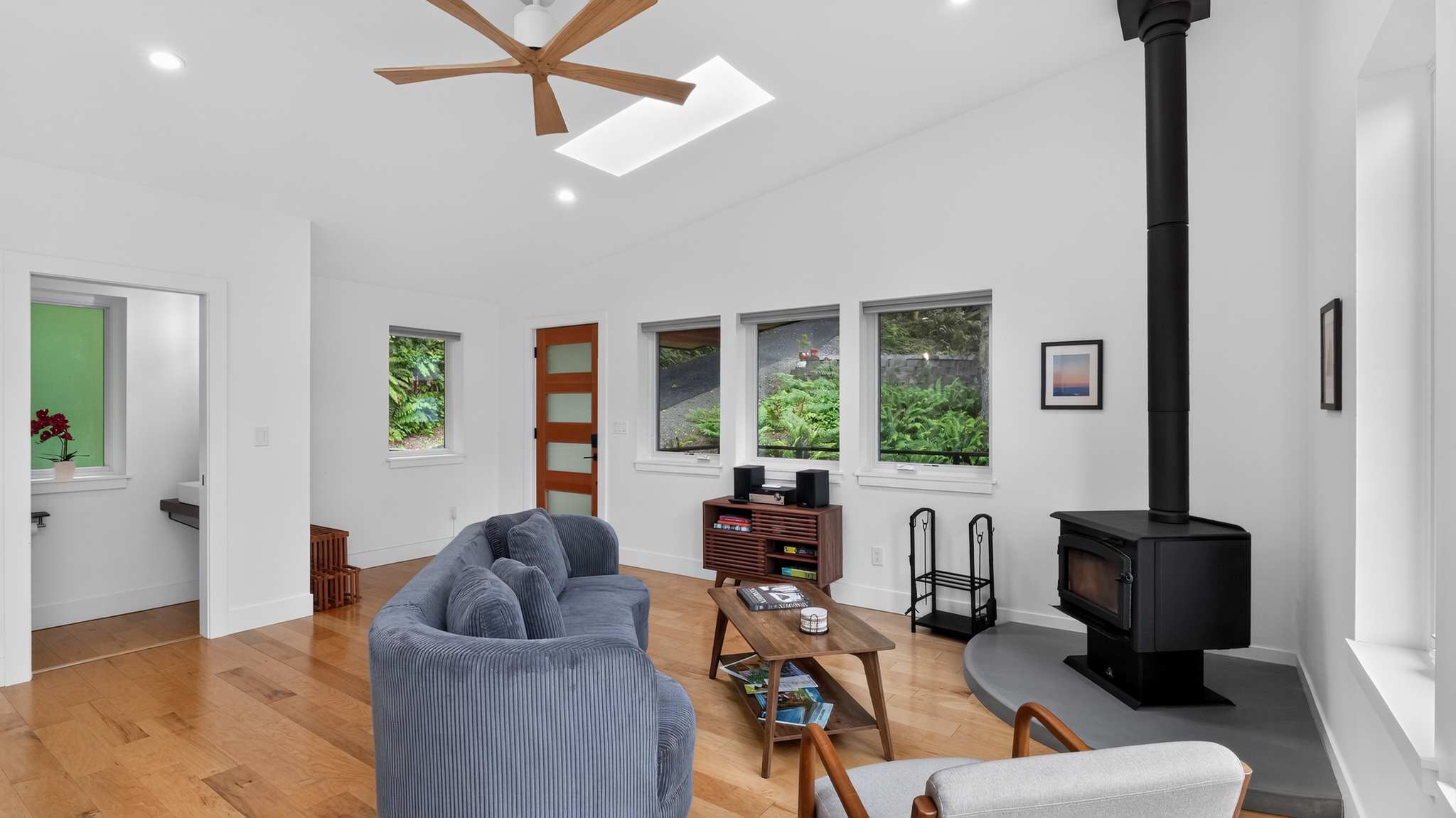 Cozy living room featuring light hardwood floors, a curved gray sofa, a black wood-burning stove on a gray hearth, and a wooden coffee table. Multiple windows provide views of greenery, and a wooden ceiling fan is overhead.