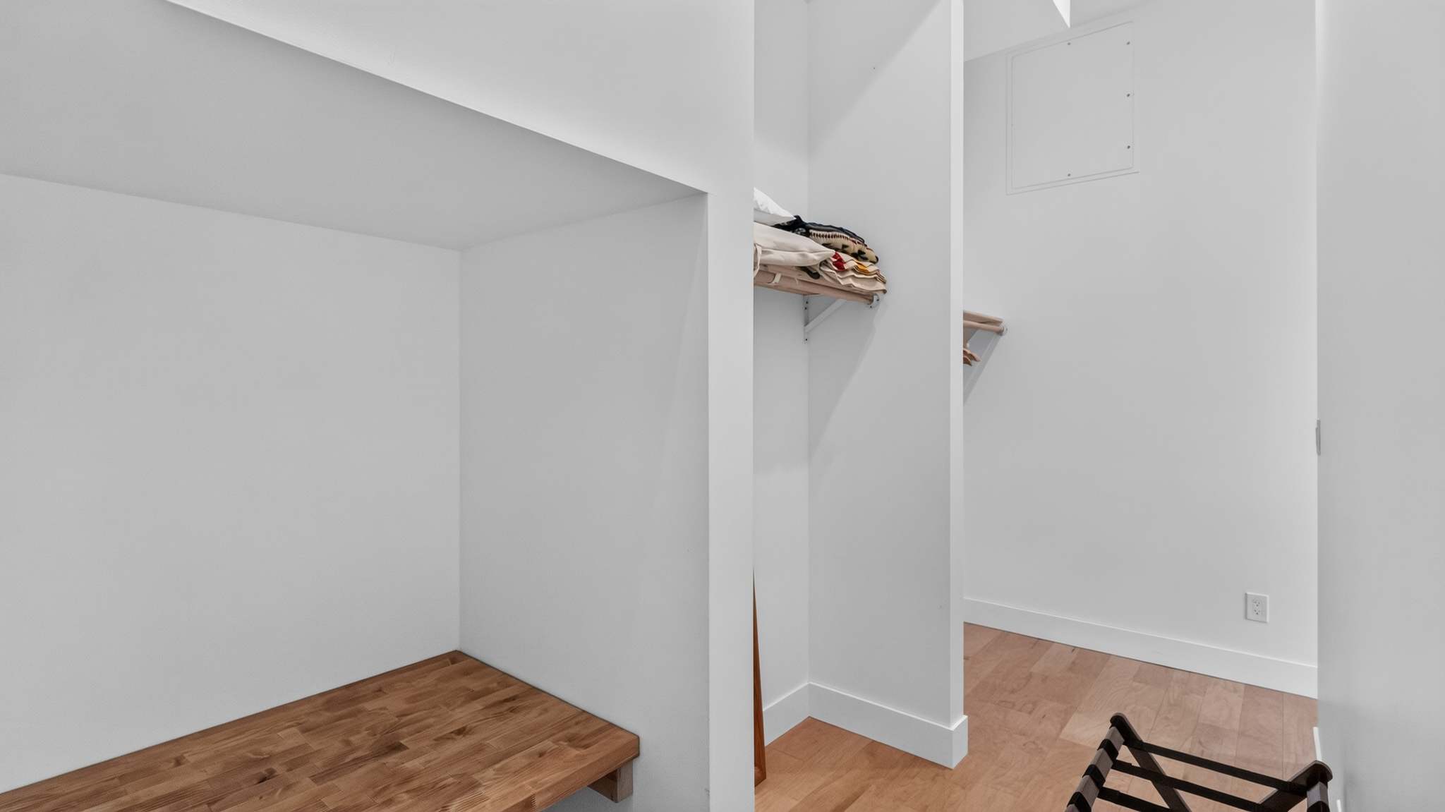 A bright white walk-in closet features a built-in wooden bench, open shelving with folded items, and a clothes rail, all set on light hardwood floors.