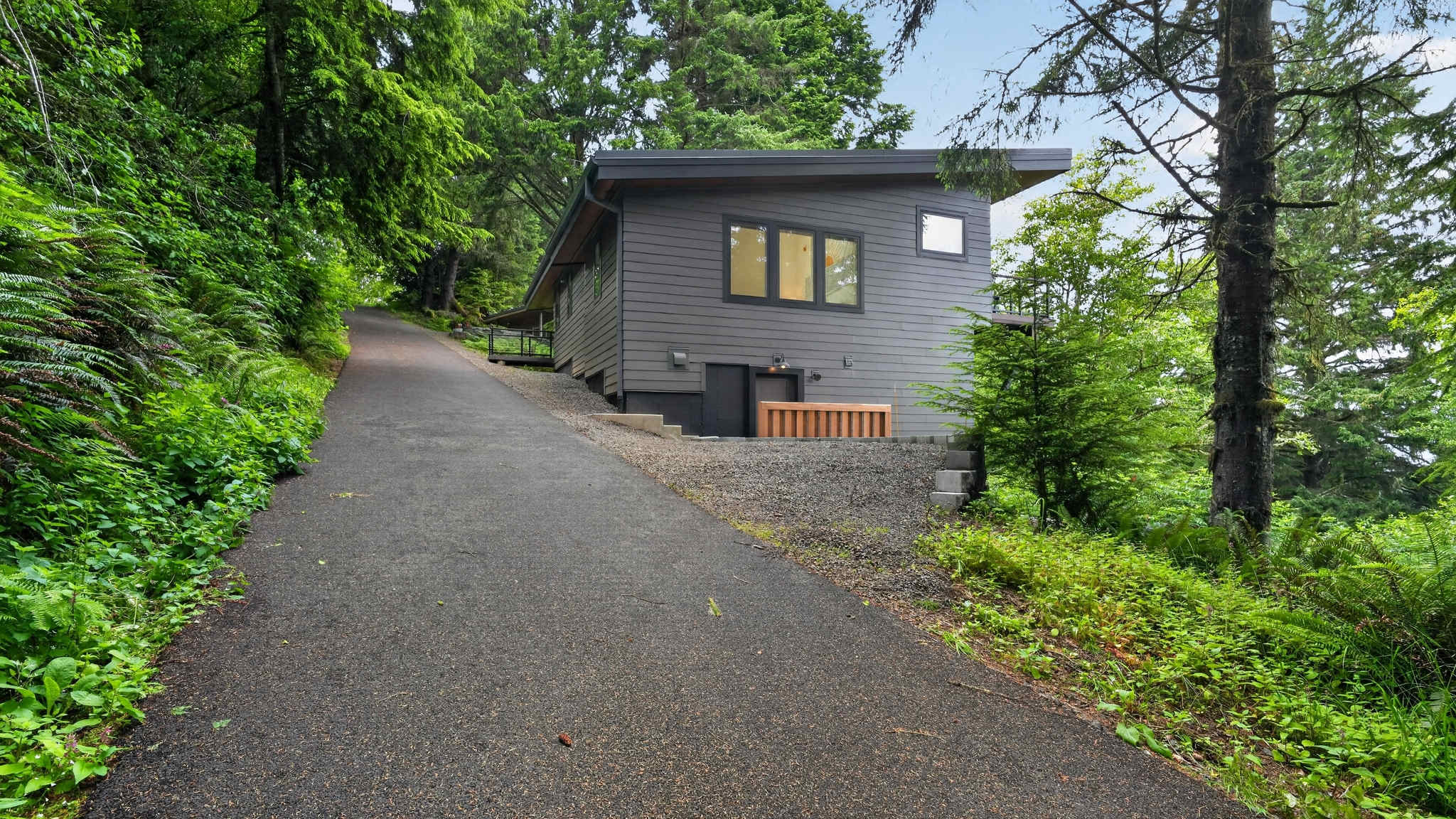 A paved driveway leads up to the side of a modern dark gray house, which is surrounded by lush green trees and foliage. A hot tub is visible on a lower level, blending into the natural landscape.