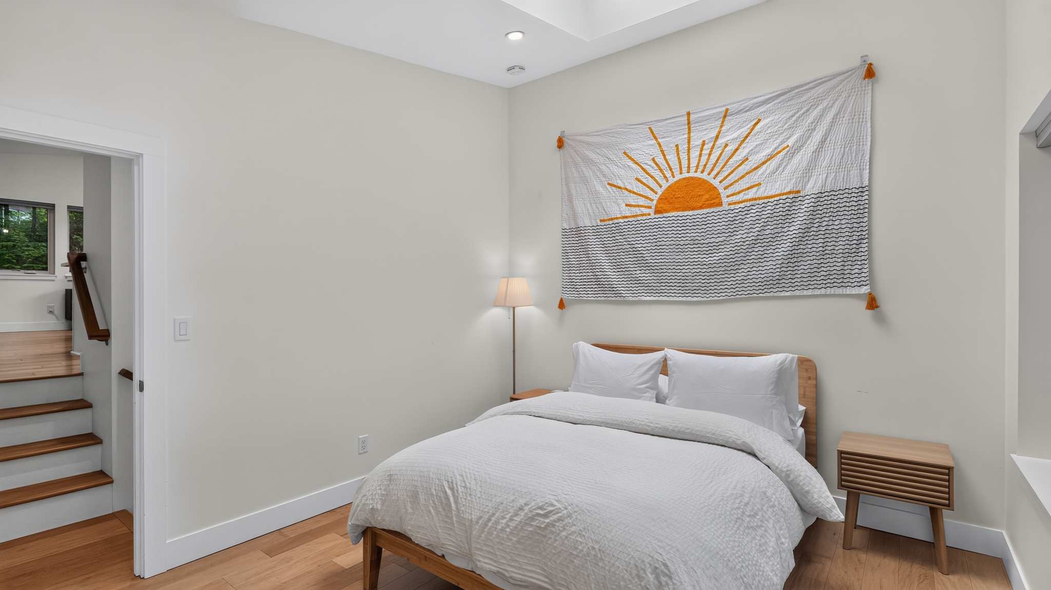Cozy bedroom with light-colored walls, a wooden bed frame, and light wood floors, featuring a large sun tapestry above the bed and a floor lamp.