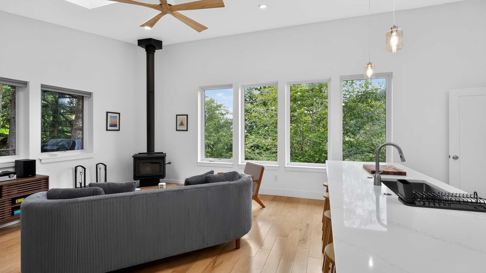 Bright, open-concept living space with white walls, light hardwood floors, a gray sofa, a black wood-burning stove, and large windows offering lush green tree views. A kitchen island with a sink is visible on the right.