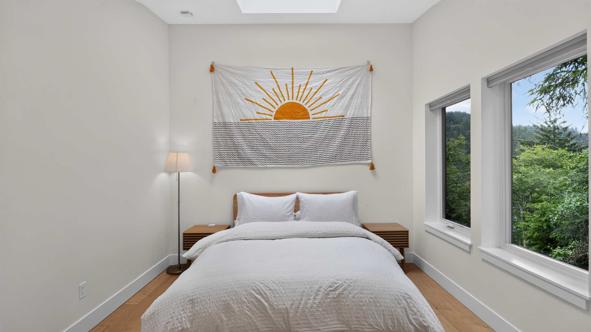 Bright bedroom with light walls, a wooden bed, and light wood floors, featuring a sun tapestry, a floor lamp, and a large window offering lush tree views.