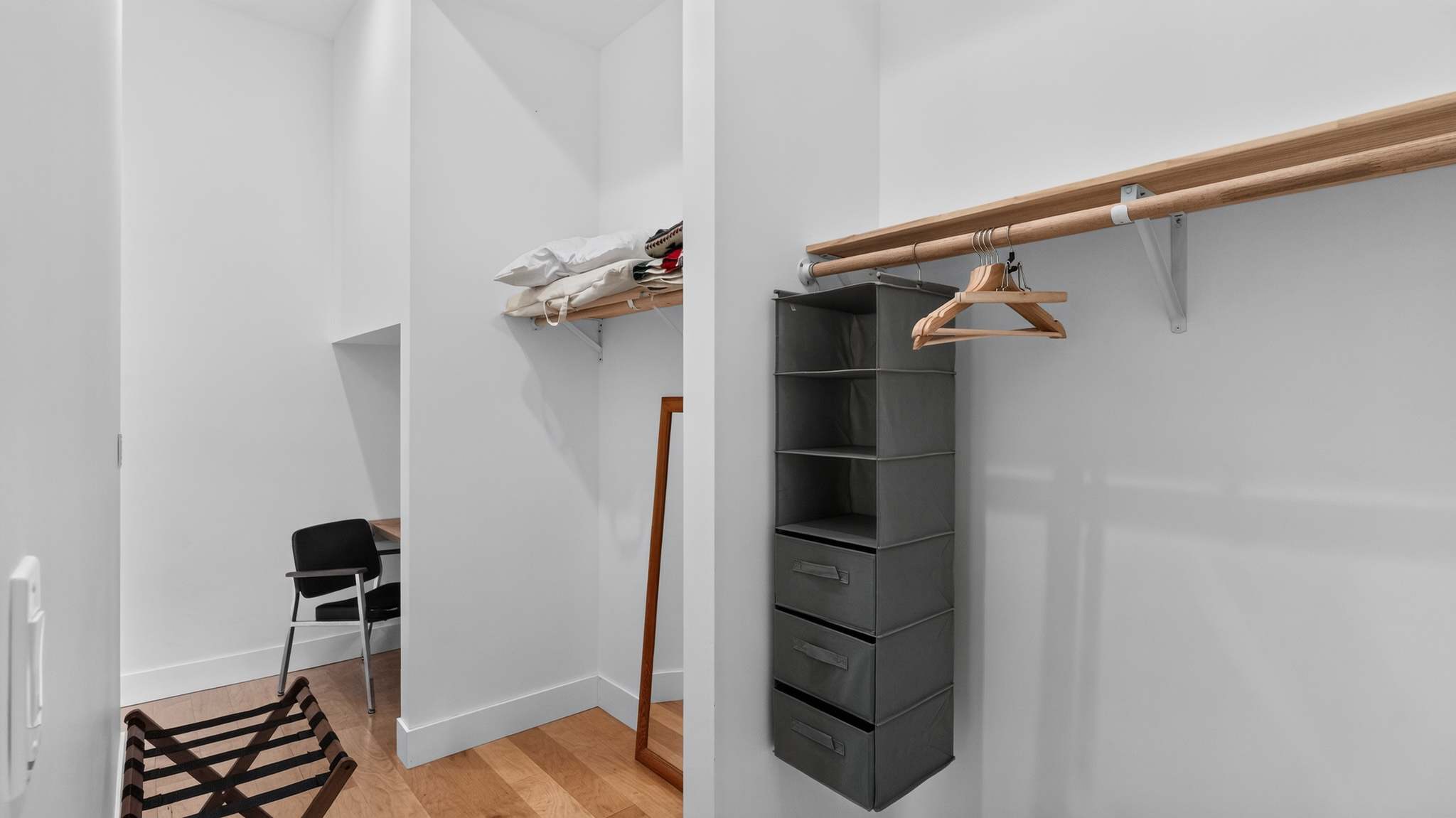 Walk-in closet with light hardwood floors and white walls, featuring a wooden hanging rod and a fabric hanging organizer with drawers. A mirror and a small desk area are also visible within the closet space.