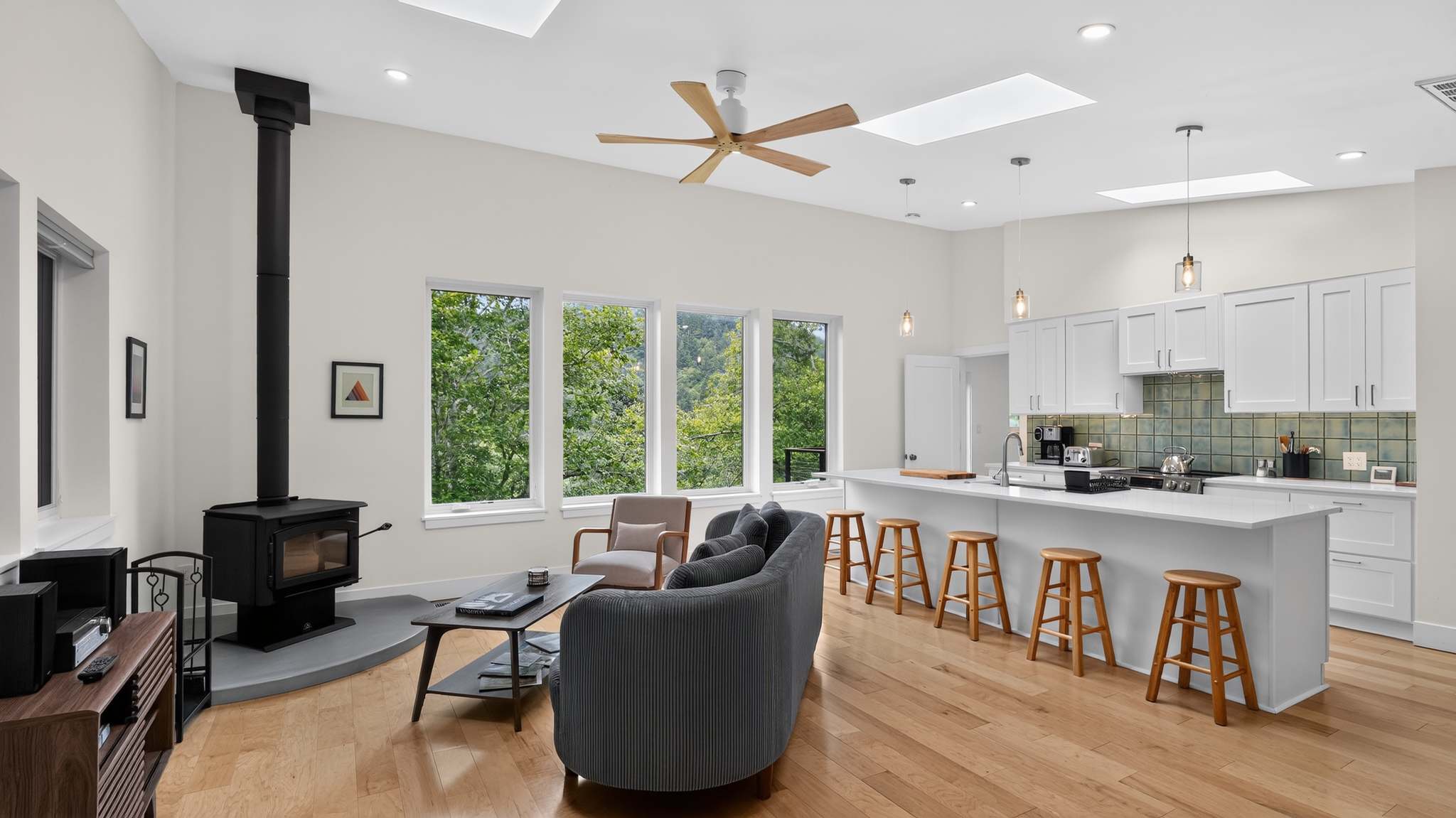 Open-concept living room with a wood-burning stove, a gray curved sofa, and large windows offering views of lush green trees, connected to a kitchen with a white island and bar stools.