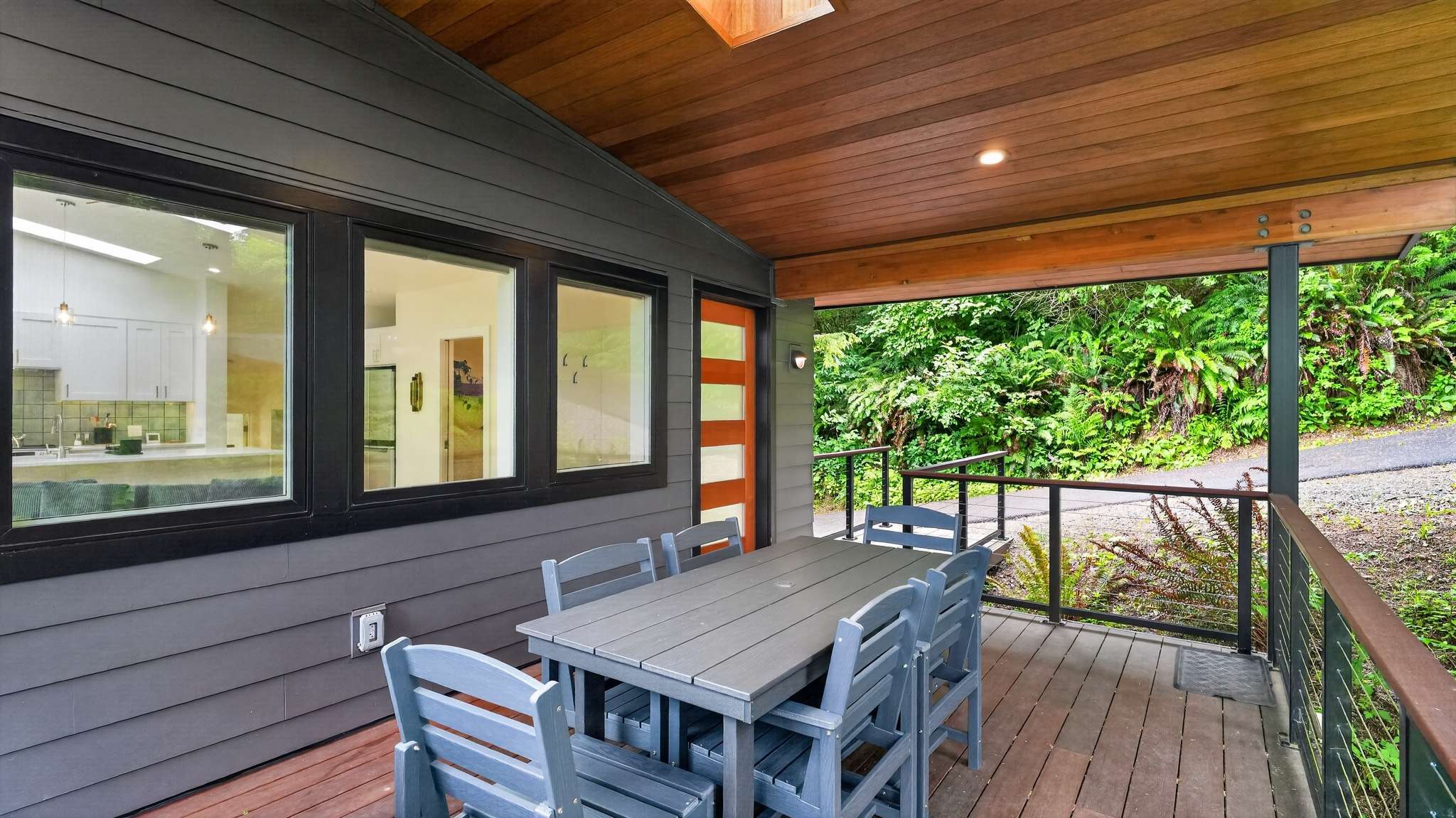 A covered outdoor deck with a grey wooden dining set and a view of the house's grey siding and large windows. A distinctive red front door with frosted glass panels is visible, leading into the home, surrounded by lush greenery.